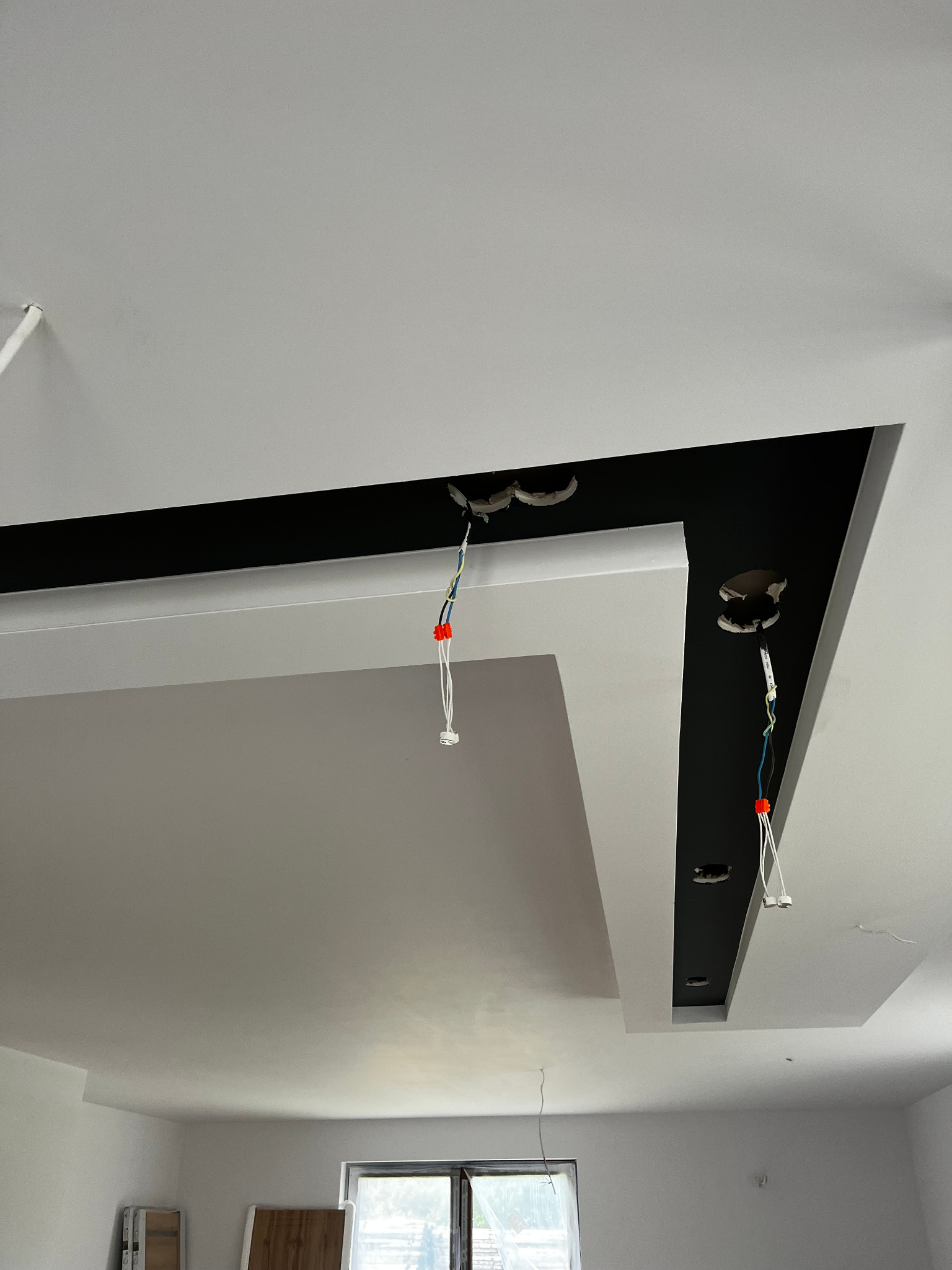 Wykończony sufit podwieszany z czarną wnęką i przygotowanymi punktami oświetleniowymi. Widoczne przewody elektryczne. W tle okno i elementy wyposażenia wnętrza.