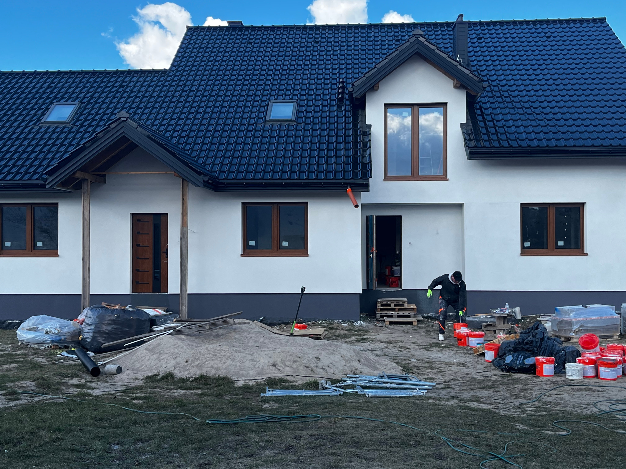 Nowo wybudowany dom z ciemną dachówką, białą elewacją i drewnianymi oknami. Na zewnątrz widoczny pracownik i materiały budowlane, sugerujące trwające prace wykończeniowe.
