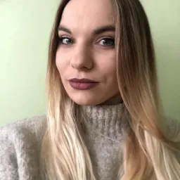 Portret kobiety z ombre włosami w szarym swetrze na tle zielonej ściany, z makijażem z akcentem na ciemną szminkę.