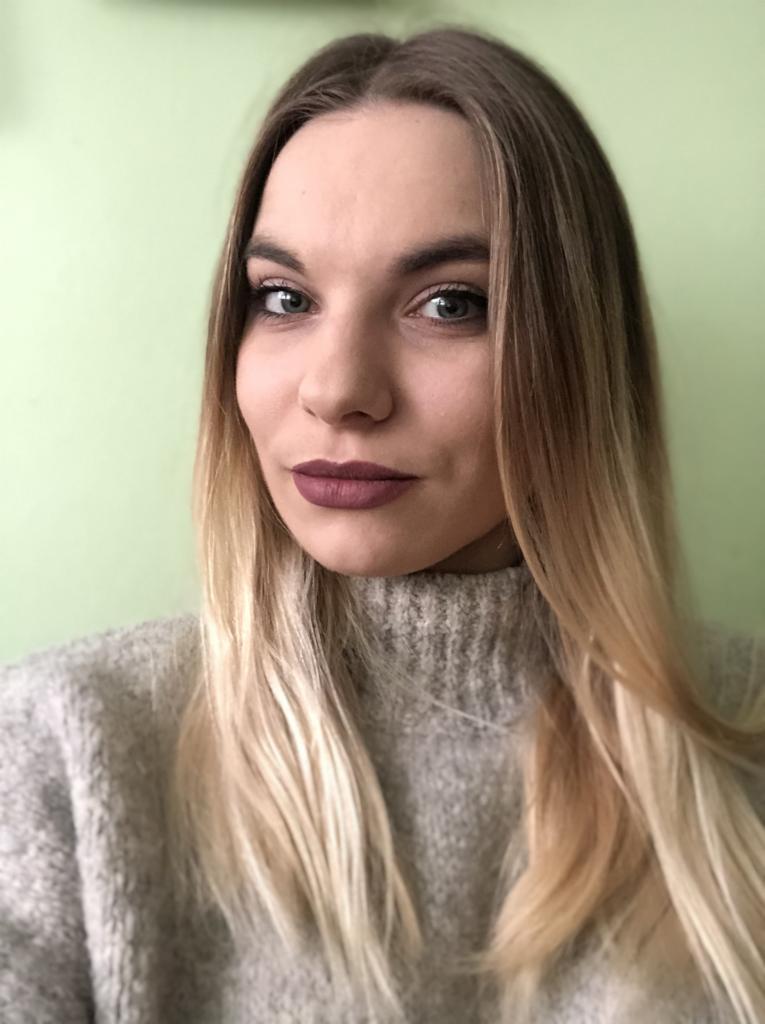 Portret kobiety z ombre włosami w szarym swetrze na tle zielonej ściany, z makijażem z akcentem na ciemną szminkę.