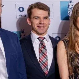 Portret uśmiechniętego mężczyzny w granatowym garniturze i kraciastej czerwono-szarej koszuli na tle ściany z logo.