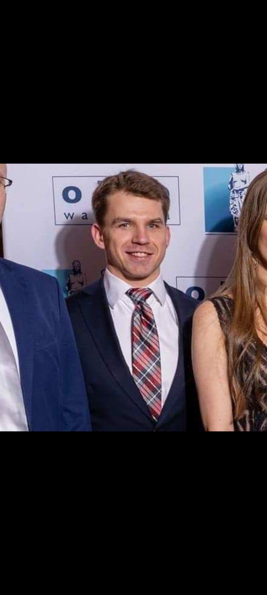 Portret uśmiechniętego mężczyzny w granatowym garniturze i kraciastej czerwono-szarej koszuli na tle ściany z logo.