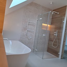 HOUSE-Technic Usługi Ogólnobudowlane - Nowoczesna łazienka z wolnostojącą wanną, kabiną prysznicową ze szklanymi drzwiami i grzejnikiem drabinkowym na ścianie z wzorem liści. Jasne płytki i oświetlenie.