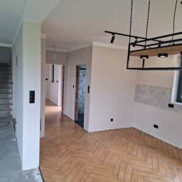 HOUSE-Technic Usługi Ogólnobudowlane - Jasne wnętrze po remoncie: parkiet w jodełkę, świeżo pomalowane ściany w odcieniach bieli i szarości, nowoczesne oświetlenie. Widok na schody i inne pomieszczenia.