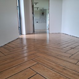 HOUSE-Technic Usługi Ogólnobudowlane - Ułożona podłoga z płytek imitujących drewno w jodełkę, widok z niskiej perspektywy. Wykończenie wnętrza w Jodłowniku. Białe ściany, widoczne przejście do innego pomieszczenia.