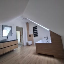 HOUSE-Technic Usługi Ogólnobudowlane - Nowoczesna łazienka na poddaszu z drewnopodobną podłogą i zabudową wanny. Umywalka z szafką i lustrem, toaleta podwieszana, grzejnik drabinkowy. Widoczne elementy wykończeniowe.