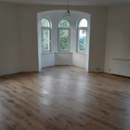 Jasne, nowo ułożone panele podłogowe z widocznymi słojami drewna w pustym pomieszczeniu z trzema oknami w kształcie łuku i świeżo pomalowanymi ścianami na biało.