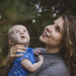 Matka z długimi brązowymi włosami trzyma niemowlę w niebieskiej koszuli, oboje patrzą w górę na tle zieleni.