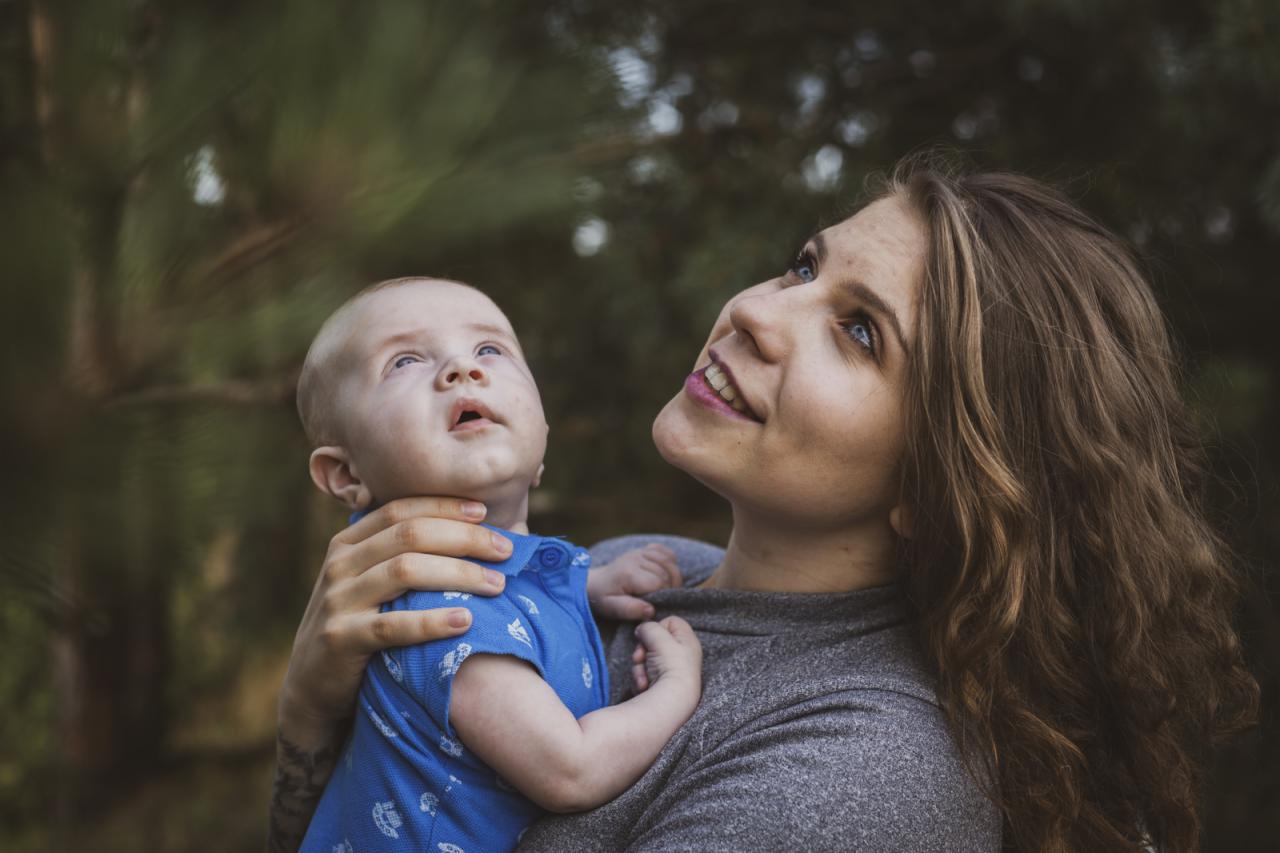 Matka z długimi brązowymi włosami trzyma niemowlę w niebieskiej koszuli, oboje patrzą w górę na tle zieleni.