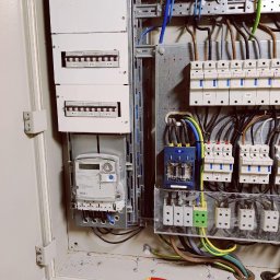 EURO-INWEST SPÓŁKA Z OGRANICZONĄ ODPOWIEDZIALNOŚCIĄ - Otwarta skrzynka elektryczna z bezpiecznikami, licznikiem i okablowaniem. Widoczne elementy instalacji elektrycznej wewnątrz obudowy.