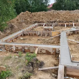 Fundamenty domu w trakcie wylewania betonu, drewniane szalunki z metalowymi łącznikami, widoczny pryzmat ziemi w tle.