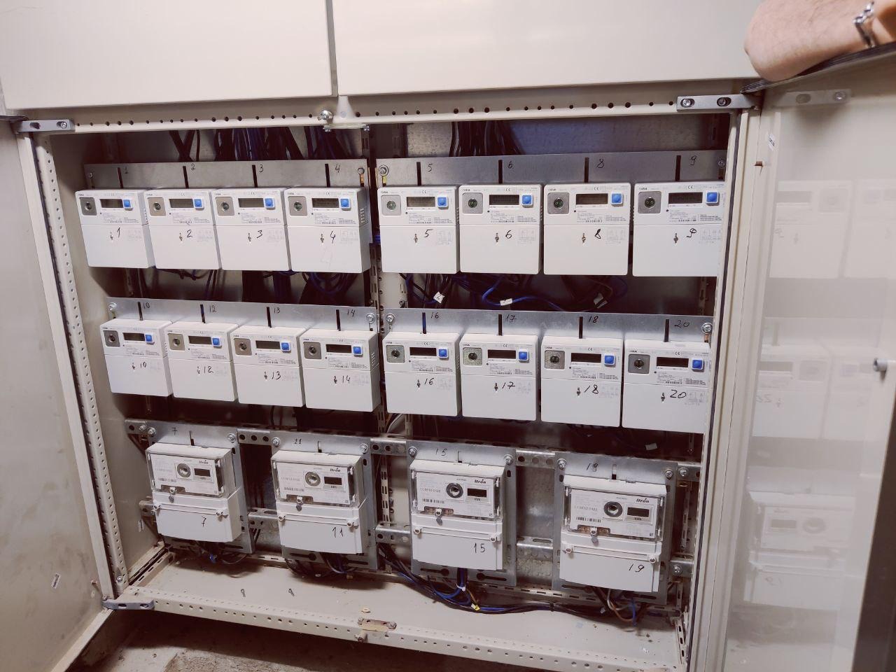 Rozdzielnia elektryczna z wieloma licznikami energii, każdy oznaczony numerem. Widoczne przewody i elementy montażowe wewnątrz metalowej szafy.