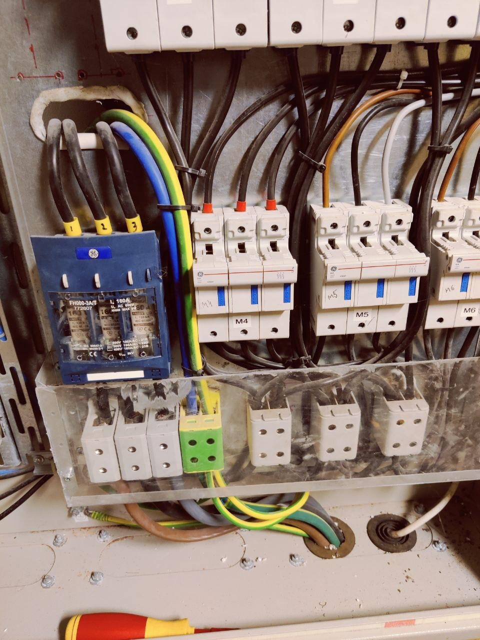 Szczegółowe ujęcie tablicy rozdzielczej z bezpiecznikami, przewodami oznaczonymi numerami i kolorami, oraz fragmentem żółto-czerwonego śrubokręta w dolnej części kadru.