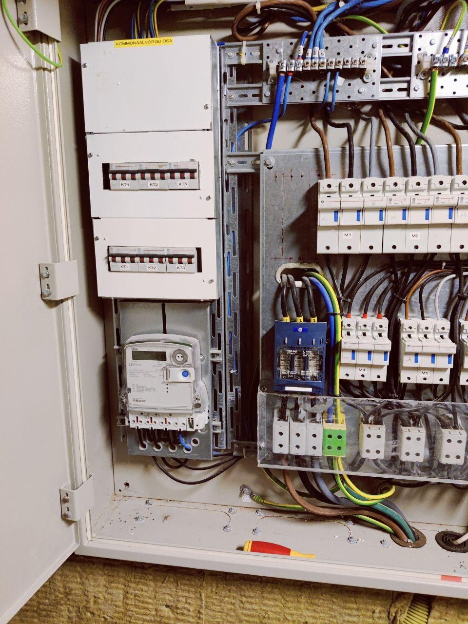 Otwarta skrzynka elektryczna z bezpiecznikami, licznikiem i okablowaniem. Widoczne elementy instalacji elektrycznej wewnątrz obudowy.