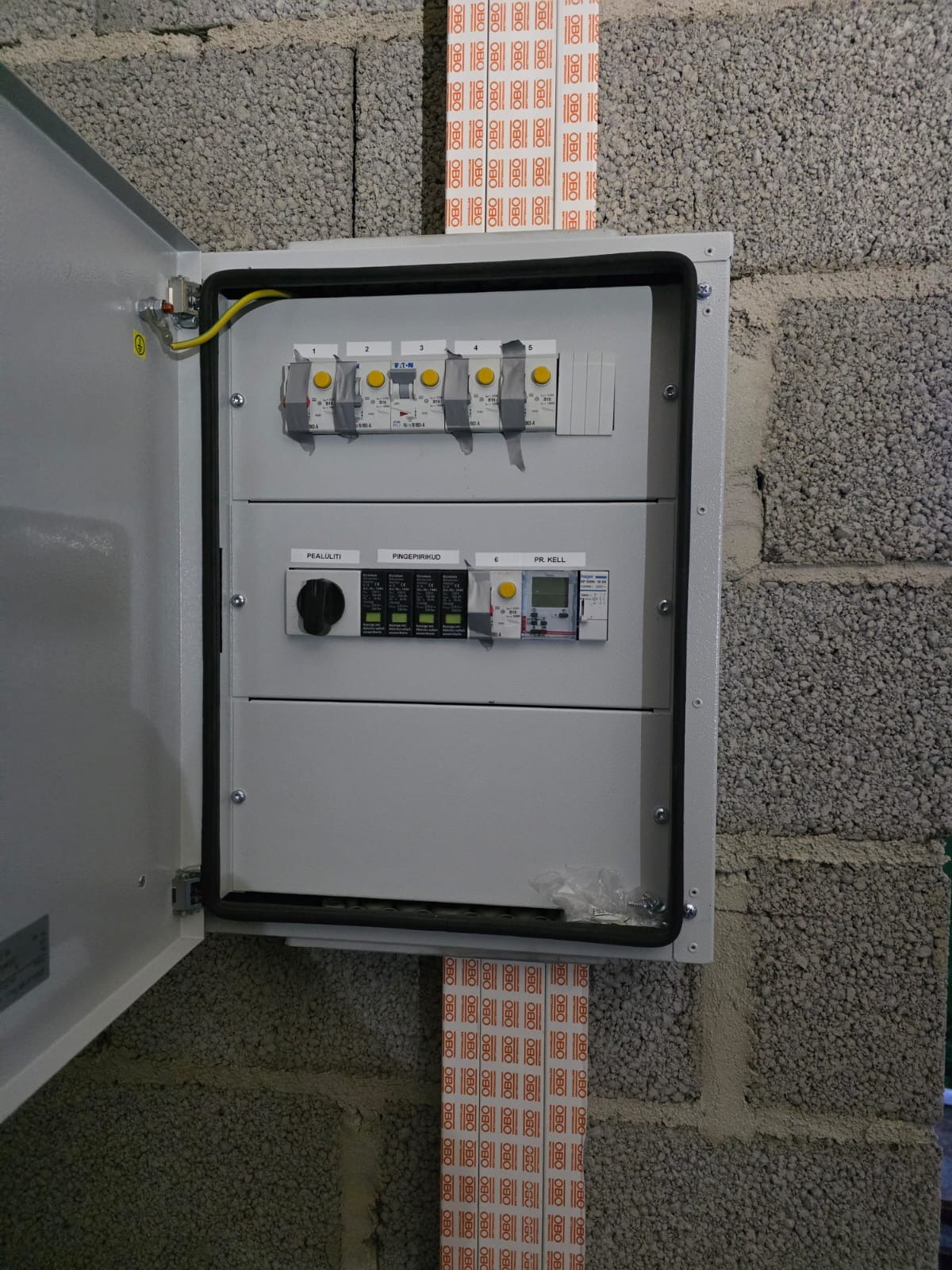 Otwarta skrzynka elektryczna z bezpiecznikami i wyłącznikami, zamontowana na szarej ścianie z bloczków betonowych. Widoczne przewody i oznaczenia na urządzeniach.