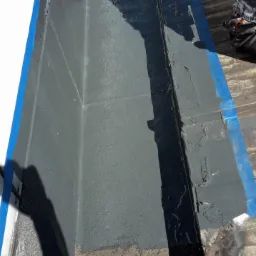 Szczegółowe ujęcie świeżo pomalowanej, szarej powierzchni dachu, zabezpieczonej niebieską taśmą malarską, z widocznymi śladami wcześniejszych prac i fragmentem sylwetki osoby w tle.