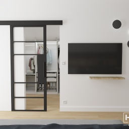 Home Effect - Minimalistyczna sypialnia z przesuwnymi drzwiami w czarnej ramie, prowadzącymi do garderoby z oświetleniem LED, na ścianie telewizor i trzy okrągłe lampy.