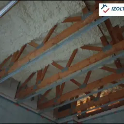 Poddasze z drewnianą konstrukcją więźby dachowej i metalowymi profilami, ocieplone białą pianką PUR z widocznym logo firmy IZOLTECH w prawym górnym rogu.