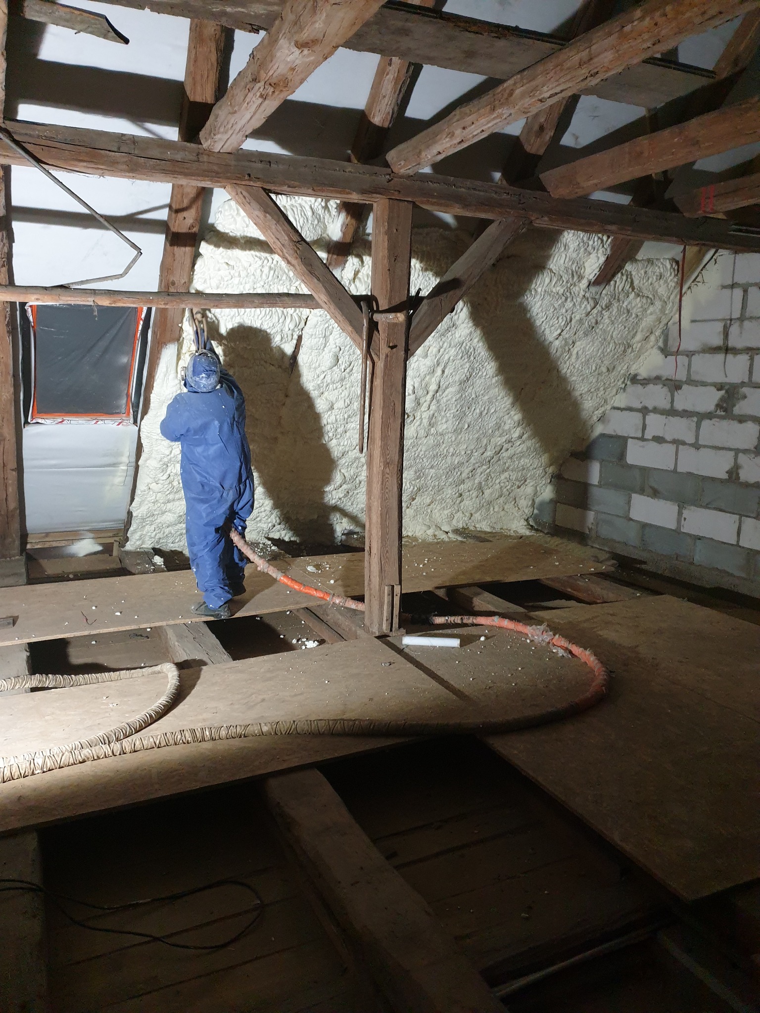 Pracownik w kombinezonie ochronnym aplikuje białą pianę izolacyjną na poddaszu z drewnianą konstrukcją więźby dachowej i fragmentami muru z bloczków.