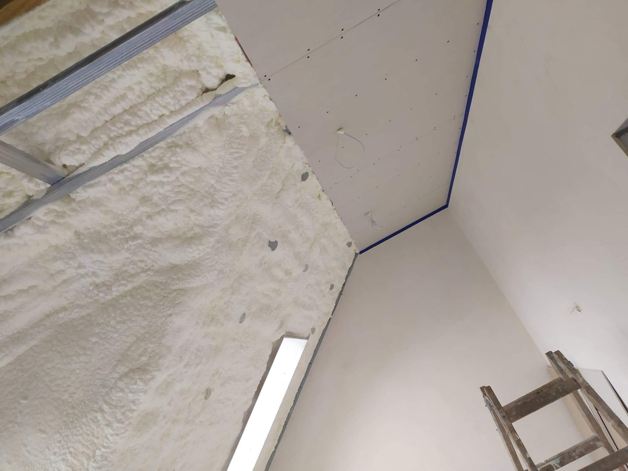 Poddasze w trakcie ocieplania: widoczna warstwa białej pianki izolacyjnej na skośnej ścianie, metalowy stelaż konstrukcyjny, fragment płyty kartonowo-gipsowej, oraz drewniana drabina oparta o ścianę.