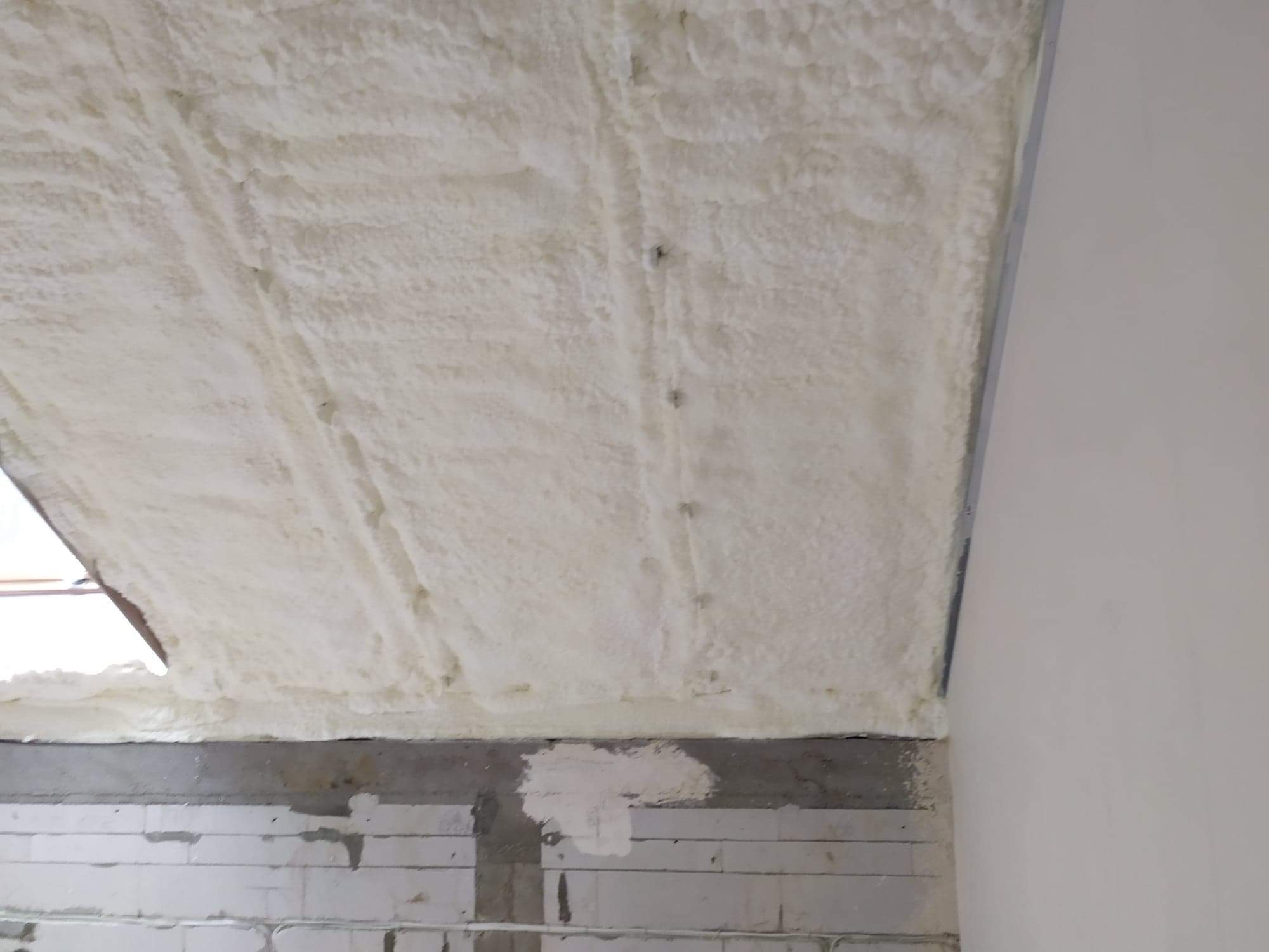 Poddasze ocieplone pianką poliuretanową, widoczne nierówności struktury piany i metalowe elementy mocujące, ściana z bloczków betonowych w dolnej części kadru.