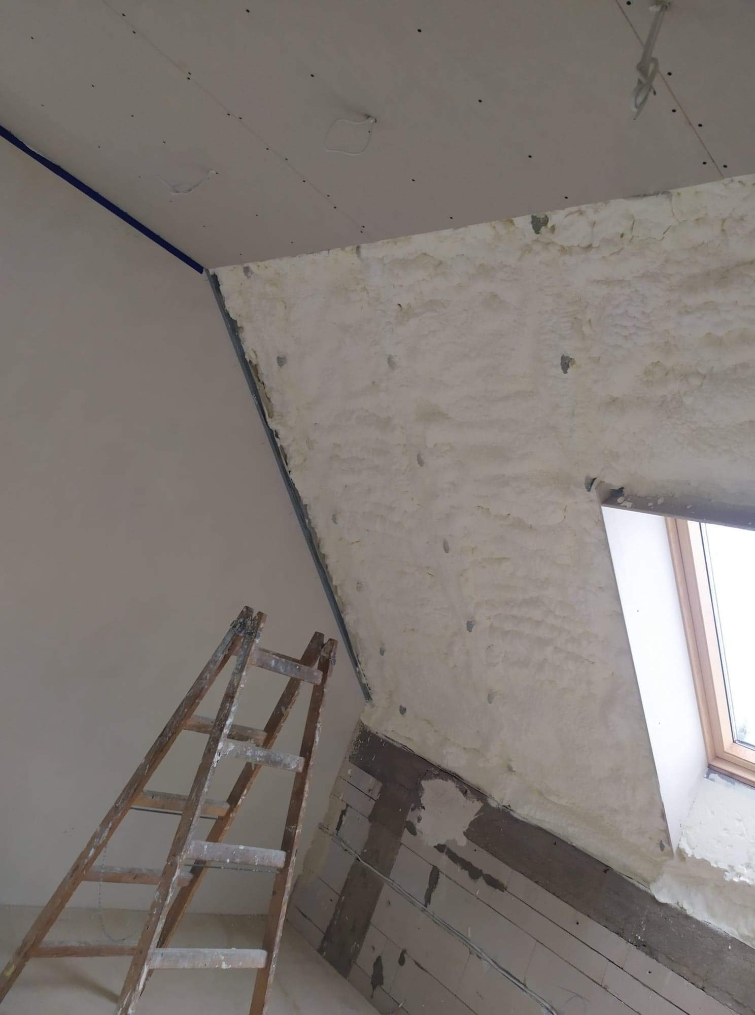 Poddasze w trakcie ocieplania: ściana pokryta pianką izolacyjną, drewniana drabina oparta o ścianę, fragment okna dachowego widoczny w rogu, sufit z płyt gipsowo-kartonowych z widocznymi śladami po...