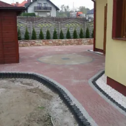 Czerwona kostka brukowa z dekoracyjnym okręgiem i obrzeżami. Widoczny fragment budynku i drewniana altana. Tło: ogrodzenie z kamienia i tujami.