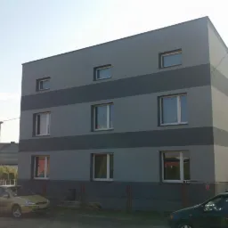 Budynek mieszkalny z szarą elewacją i pasami ciemniejszego szarego tynku. Widoczne okna z białymi ramami i zaparkowane samochody.