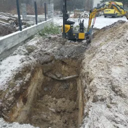 Żółta mini koparka wykonuje wykop w ziemi pokrytej lekkim śniegiem, widoczny fragment instalacji w wykopie, w tle ogrodzenie z kostki i zaparkowane samochody dostawcze.