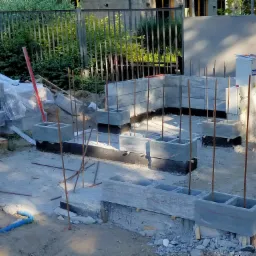 Wznoszenie fundamentów z betonowych bloczków z pustymi przestrzeniami, wystające pręty zbrojeniowe, widoczne zarysy przyszłych ścian, teren budowy z gruzem i narzędziami.