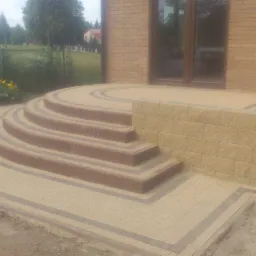 Półokrągłe schody z kostki brukowej prowadzące do wejścia domu, z obrzeżami w kontrastowym kolorze i niskim murkiem z boku.