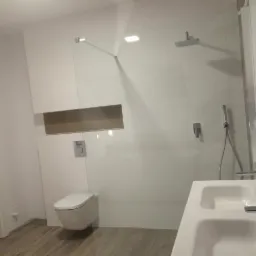 Nowoczesna łazienka z białą armaturą, podwójną umywalką, minimalistyczną toaletą podwieszaną i przeszklonym prysznicem typu walk-in z deszczownicą, wykończona płytkami w odcieniach bieli i szarości.