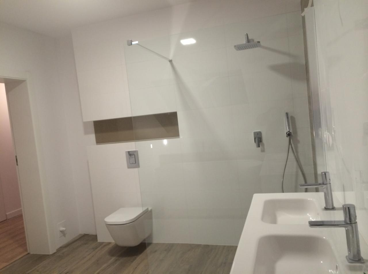 Nowoczesna łazienka z białą armaturą, podwójną umywalką, minimalistyczną toaletą podwieszaną i przeszklonym prysznicem typu walk-in z deszczownicą, wykończona płytkami w odcieniach bieli i szarości.