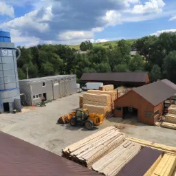 Widok z góry na teren tartaku z placem składowym, stosem tarcicy, maszyną załadunkową i budynkami przemysłowymi na tle zielonego krajobrazu.