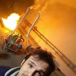 Mężczyzna robi selfie na tle urządzenia do symulacji pożaru z płomieniami i dymem, stojącego na wózku z butlami. Widoczne rury odprowadzające dym.