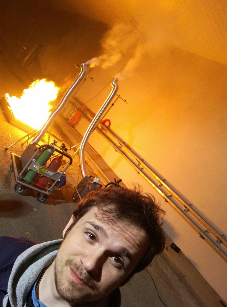 Mężczyzna robi selfie na tle urządzenia do symulacji pożaru z płomieniami i dymem, stojącego na wózku z butlami. Widoczne rury odprowadzające dym.