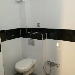 Wykończona łazienka z białymi i czarnymi płytkami ułożonymi w pasy, wisząca toaleta z chromowanym przyciskiem spłukiwania, szara podłoga imitująca drewno, dodatkowy bidet podłączony do baterii.