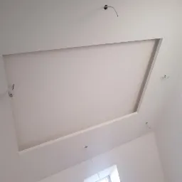 Wykończony sufit podwieszany z prostokątnym wnęką, widoczne wystające przewody elektryczne i fragment okna z niebieską ramą.