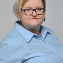 Portret kobiety w okularach i błękitnej koszuli na szarym tle.