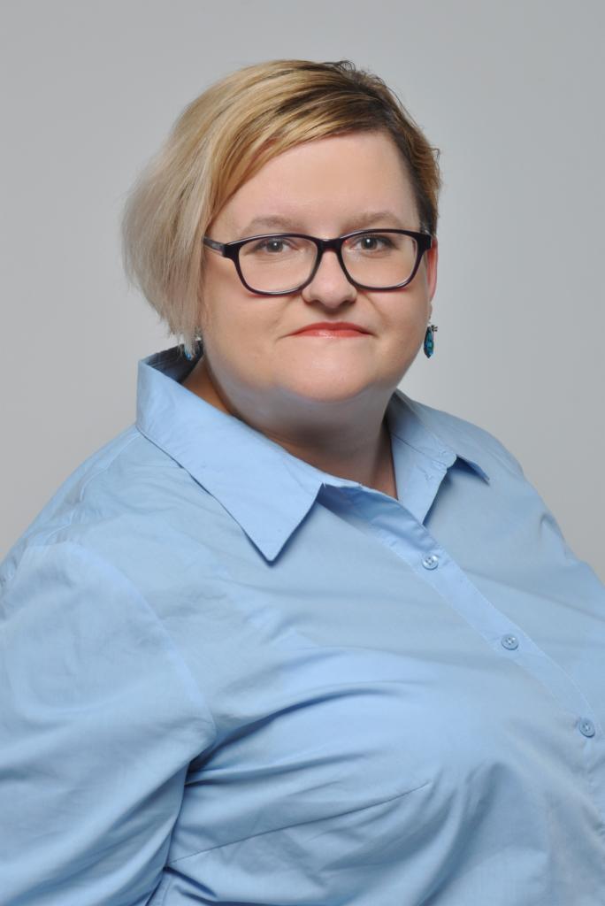 Portret kobiety w okularach i błękitnej koszuli na szarym tle.