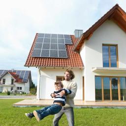 Sunpark Polska Sp. z o.o. - Uśmiechnięty ojciec podnosi syna na tle nowoczesnego domu z panelami słonecznymi na dachu i trawnikiem.