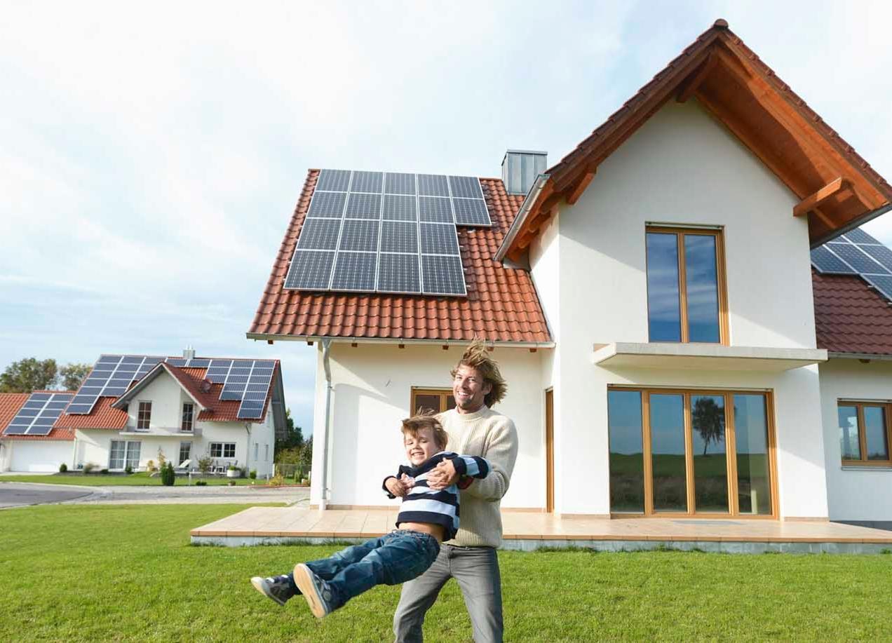 Uśmiechnięty ojciec podnosi syna na tle nowoczesnego domu z panelami słonecznymi na dachu i trawnikiem.