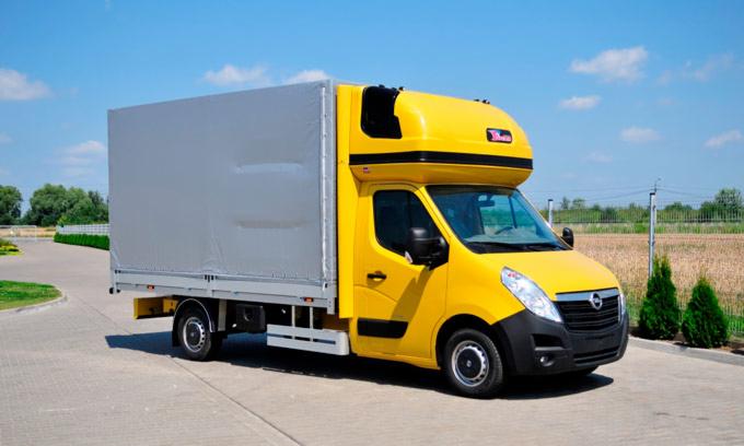 Żółty samochód dostawczy z zabudową plandekową na tle błękitnego nieba i zieleni.