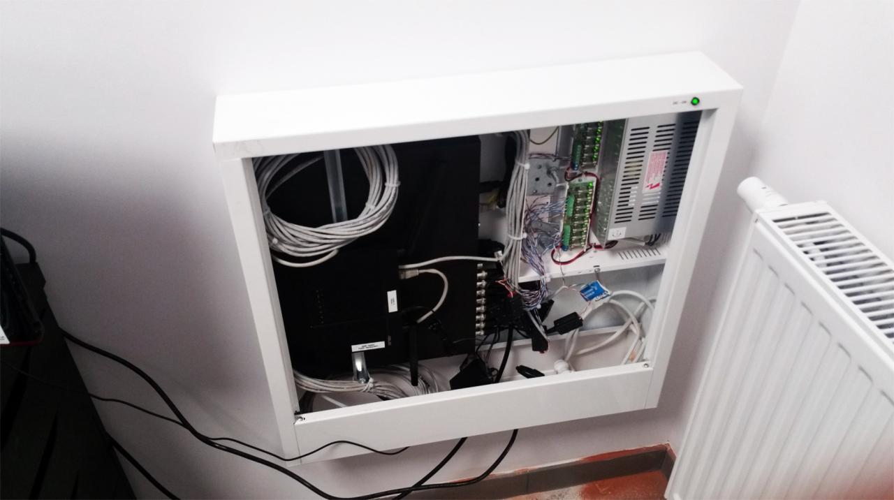 Otwarta, biała szafka telekomunikacyjna zamontowana na ścianie, z widocznymi kablami, złączami i elementami elektroniki wewnątrz, obok grzejnik.