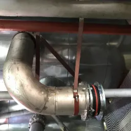 Metalowy, zakrzywiony przewód rurowy z kompensatorem, zamocowany do konstrukcji wsporczej w hali przemysłowej.