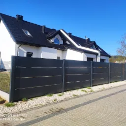 Nowoczesne, antracytowe ogrodzenie panelowe z poziomymi listwami, oddzielające posesję z białym domem o czarnym dachu od chodnika z kostki brukowej.