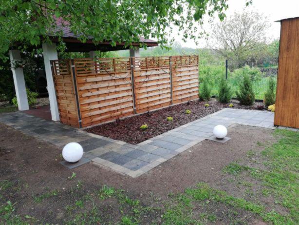 Ułożona kostka brukowa tworząca ścieżkę obok drewnianego płotu z ozdobnym wykończeniem, oświetlona kulistymi lampami, z rabatą wysypaną korą i niskimi roślinami iglastymi w tle.