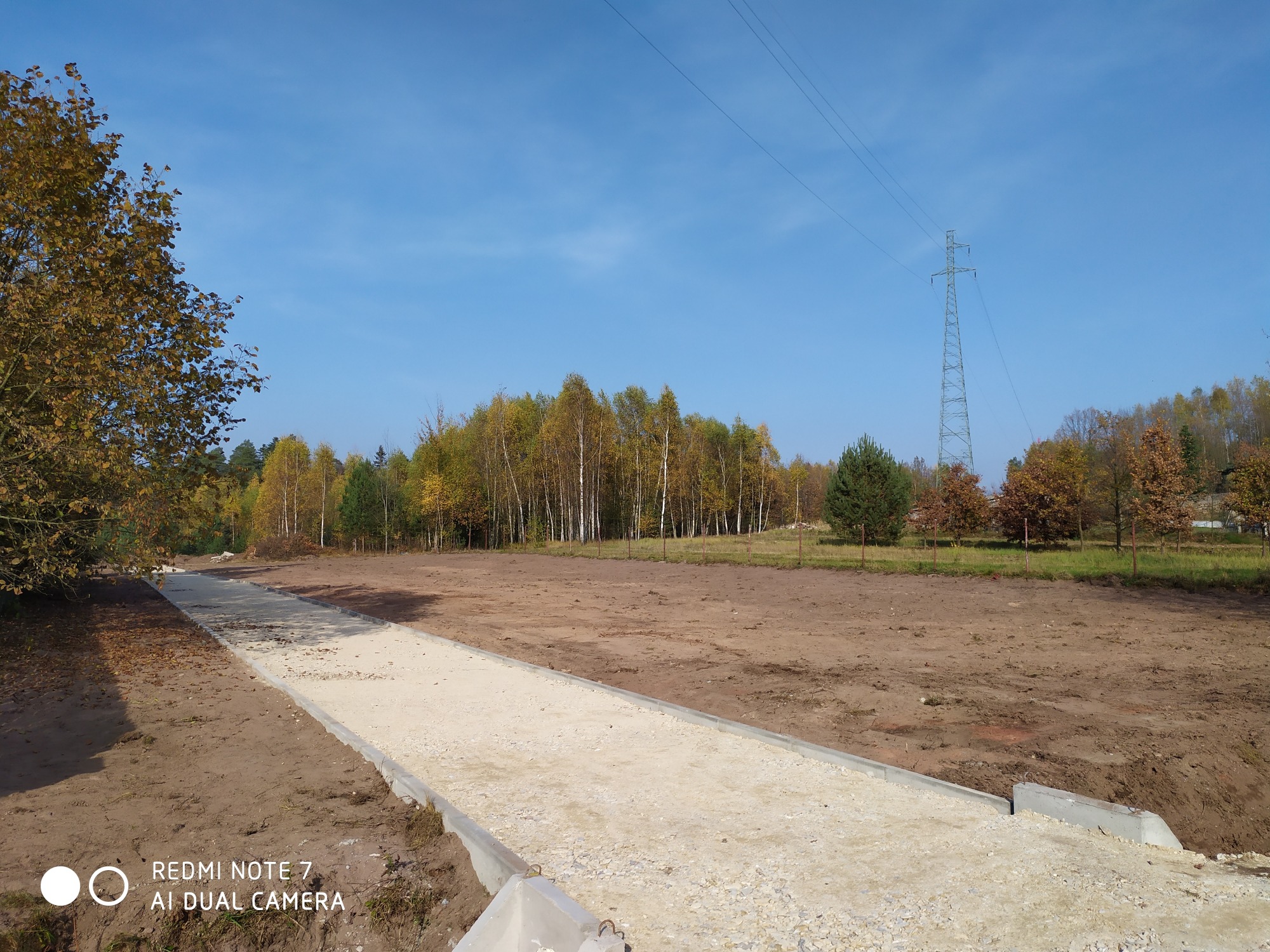 Wysypana żwirem droga w terenie z przygotowanym podłożem, widoczne betonowe obrzeża, w tle drzewa i słup wysokiego napięcia pod błękitnym niebem.
