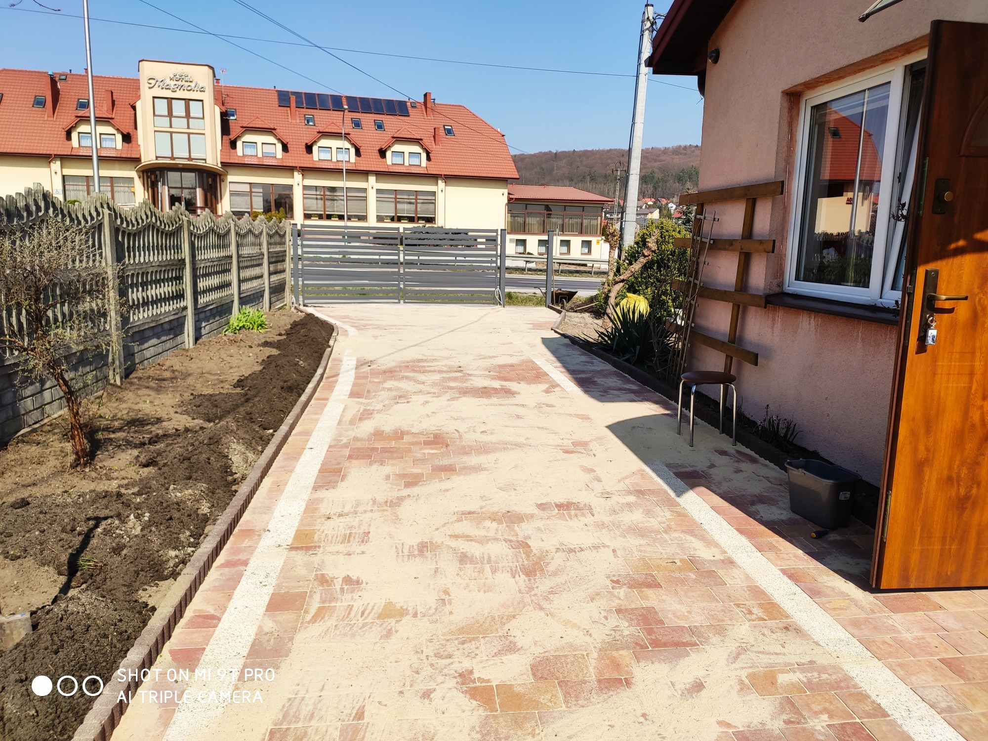 Nowa nawierzchnia z ceglanej kostki prowadząca do budynku, obrzeża z jasnego kruszywa, przygotowane rabaty po bokach, widoczny budynek w tle.