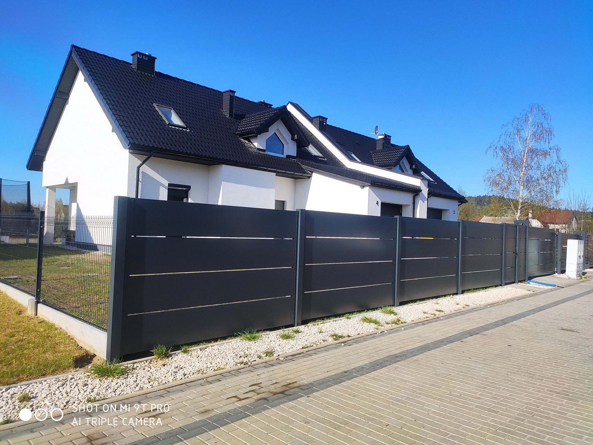 Nowoczesne, antracytowe ogrodzenie panelowe z poziomymi listwami, oddzielające posesję z białym domem o czarnym dachu od chodnika z kostki brukowej.